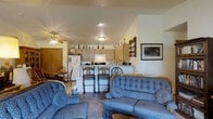 This single-floor residence showcases an open-concept design that integrates the living, dining, and kitchen areas into one cohesive, flowing space. The vaulted ceiling adds a sense of vertical spaciousness, enhanced by a ceiling fan that provides both function and a casual aesthetic.

The living area is furnished with a coordinated blue upholstered sofa set, including a loveseat and armchair, arranged to encourage comfortable conversation. A framed landscape print adorns the wall, adding a decorative focal point to the space.

The kitchen features light wood cabinetry, a white refrigerator, and a range with an overhead hood. A breakfast bar with two bar-height chairs provides a casual dining option and acts as a natural divider between the kitchen and living areas. Open shelving and a wine rack add character and practical storage.

Adjacent to the kitchen, a dining area accommodates a wooden table and chairs, offering a more formal setting for meals. The space also includes a large wooden armoire-style cabinet, adding substantial storage. Overall, the home presents a warm, functional, and cohesive single-level living environment.