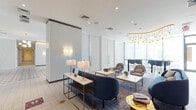 This single-floor amenity space is thoughtfully designed with a cohesive contemporary aesthetic, blending refined materials, curated furnishings, and abundant natural light throughout. The main lobby and lounge areas feature distinctive patterned encaustic tile flooring with floral and geometric motifs, complemented by plush navy and dark plum upholstered seating arranged to encourage relaxed gatherings. Slatted white wall paneling adds texture and depth, while globe chandeliers and brass wall sconces provide warm, layered illumination. Floor-to-ceiling sheer-draped windows invite generous daylight and connect the interior to surrounding outdoor greenery. A dedicated lounge zone transitions into an elevated bar-height communal table area, anchored by a striking sculptural chandelier with cascading brass arms and globe pendants. A glass-partitioned office or leasing area offers a more private setting, appointed with light wood flooring, a teal sofa, and gold-accented task lighting. The game room provides a vibrant social hub complete with a billiards table, ping-pong table, built-in banquette seating, and bold graphic wall art with gold pendant lighting. Throughout the space, brass hardware and gold accents provide a consistent thread of understated luxury.