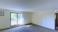 This single-floor residential space offers a clean and functional layout designed for comfortable everyday living. The main living area is the centerpiece of the space, characterized by its open feel and neutral, light-toned walls that create a calm and welcoming atmosphere. Underfoot, soft wall-to-wall carpeting adds warmth and comfort to the room.

A standout feature of the living area is the large sliding glass door that floods the space with abundant natural light, casting bright patches across the floor and lending an airy quality to the interior. The sliding door opens to an outdoor balcony, framed by a wooden railing and surrounded by lush greenery, providing a pleasant outdoor retreat with scenic views of the surrounding landscape.

A baseboard heating unit runs beneath the window, ensuring efficient climate control throughout the cooler months. The ceiling features a classic textured finish, adding subtle character to the otherwise clean, minimalist aesthetic of the space. Recessed ventilation is also present, contributing to overall comfort and air circulation.

The overall ambiance of this space is bright, neutral, and move-in ready, making it well-suited for a variety of interior styles and arrangements. The connection between the interior living space and the outdoor balcony is a particularly appealing element, enhancing the sense of openness and livability.