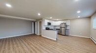 This single-floor unit offers a clean, modern layout designed for comfortable everyday living. The open-concept living and kitchen area is anchored by warm light wood-tone luxury vinyl plank flooring and recessed lighting throughout, creating a bright and welcoming atmosphere. The kitchen is well-equipped with a two-tone cabinet design — white upper shaker-style cabinets paired with gray lower cabinets — complemented by a white subway tile backsplash, dark countertops, and a full suite of stainless steel appliances including a gas range, microwave, and refrigerator. A peninsula with additional counter space separates the kitchen from the open living area, which is generously sized and flooded with recessed light. The bedrooms are finished with plush gray carpet and feature windows with horizontal blinds that allow for natural light. Baseboard heating is present throughout the unit. An additional room with vinyl plank flooring, a window air conditioning unit, and baseboard heating provides flexible living or office space. The bathroom is neatly finished in white, featuring a vanity, toilet, and wood-look flooring, with a towel bar adding a practical touch. Closet storage is accessible off the main living area, rounding out the functional design of this well-maintained unit.