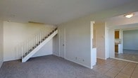 This two-story space is designed with a straightforward, functional layout that makes efficient use of its footprint. The main living area is finished with wall-to-wall carpet in a neutral gray tone, providing a comfortable and cohesive foundation for the space. A staircase with white metal railings runs along one wall, leading to the upper floor while incorporating a door beneath it that provides access to additional storage or utility space.

Adjacent to the carpeted living area, a transition to light-colored tile flooring signals a separate functional zone, likely a kitchen or dining area. This adjoining space features a pass-through or partial wall opening that connects it visually to the main area, promoting an open feel between the two zones. A closet with louvered doors is also visible in this section, offering convenient built-in storage.

The walls throughout are finished in a soft, warm beige, complementing the neutral flooring choices and contributing to a clean, understated aesthetic. The overall ambiance is calm and move-in ready, with a simple palette that provides a versatile backdrop for a variety of interior styles.