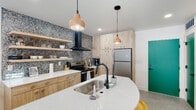 Located at 156 E 900 S in Salt Lake City, Utah, this single-floor space showcases a thoughtfully designed interior with contemporary finishes throughout. The kitchen is a standout feature, centered around a curved quartz island with an undermount sink and matte black faucet. Light wood cabinetry with rounded hardware lines the walls, complemented by open floating shelves and a dramatic terrazzo-style backsplash that extends behind the range. A wall-mounted black range hood, stainless steel appliances — including a full-size refrigerator and electric range — and pendant lighting with warm terracotta tones complete the culinary space. A vivid teal entry door adds a bold pop of color. The bedroom is bright and inviting, featuring sliding glass doors that open to a private balcony, allowing natural light to fill the room and offering views of the surrounding exterior. The bathroom continues the cohesive design language with full-height warm-toned ceramic tile, a wood-framed vanity with a white quartz countertop, a matte black faucet, an arched mirror, and a soaking tub with a curtain enclosure. Polished concrete floors run throughout, tying together the modern, nature-inspired aesthetic of this well-appointed urban space.