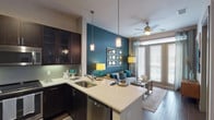 This 1-bedroom, 1-bathroom residence delivers a cohesive open-concept layout that seamlessly connects the kitchen, dining, and living areas. The kitchen is a standout feature, outfitted with rich dark cabinetry, stainless steel appliances including a French door refrigerator, built-in microwave, and range, complemented by a sleek tile backsplash and light quartz countertops. A peninsula with pendant lighting serves as both a functional workspace and casual dining area, flowing naturally into the living space. The living room makes a bold design statement with a deep teal accent wall, hardwood-style flooring, a ceiling fan, and large windows that invite generous natural light and access to an outdoor area. The bedroom is a serene retreat featuring plush carpeting, a calming blue-gray accent wall, ceiling fan, and large windows that continue the theme of natural light. The bathroom is finished with a dual-sink vanity topped with a wide mirror, and a glass-enclosed walk-in shower with elegant tile surround. Throughout the residence, neutral tones are punctuated by teal accents, creating a modern and inviting aesthetic that ties each space together cohesively.