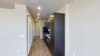 This single-floor space offers a clean, contemporary design highlighted by a well-appointed kitchen and a functional open hallway. The kitchen is outfitted with rich, dark espresso cabinetry that extends from floor to ceiling, providing ample storage space. Black appliances — including a refrigerator and gas range — complement the cabinetry, while light stone countertops offer a striking contrast. Recessed lighting throughout ensures the space remains bright and welcoming. The warm wood-look plank flooring runs continuously from the hallway into the kitchen, lending a cohesive and inviting feel underfoot. A standout architectural detail is the white sliding barn door, which adds a touch of rustic charm against the otherwise modern aesthetic. The hallway is generously proportioned, with neutral white walls and additional recessed lighting that guides the eye toward the rest of the unit. Ceiling-mounted HVAC vents are thoughtfully integrated, contributing to a tidy and uncluttered overhead aesthetic. Overall, the space balances modern finishes with practical design, creating a functional yet stylish environment suitable for everyday living.