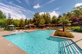 Located at 138 Greison Trail in Newnan, Georgia, this impressive outdoor amenity complex offers a resort-caliber pool experience set within beautifully maintained grounds. The centerpiece of the space is a large, multi-section swimming pool with crystal-clear turquoise water, complemented by a separate wading or spa area featuring gentle water jets. A distinctive raised island planter, anchored by a striking palm tree and overflowing with vibrant seasonal flowers, serves as a dramatic focal point within the pool area. The surrounding hardscape features spacious pool decking finished in warm-toned pavers, providing ample room for lounge chairs and sun umbrellas. Multiple covered pergola structures offer shaded gathering areas, while a gazebo with a fireplace chimney creates a cozy outdoor retreat. Stone retaining walls with integrated landscaping beds add architectural depth and a refined, layered aesthetic to the grounds. The backdrop is framed by a handsome multi-story colonial-style clubhouse building with dormer windows, white balcony railings, and brick detailing. Mature trees surrounding the perimeter provide natural privacy and a lush, green canopy. The overall ambiance is one of upscale, resort-inspired outdoor living in a well-appointed community setting.