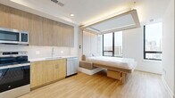 Located on the 11th floor at 4909 Auburn Ave in Bethesda, MD, this efficiently designed studio unit makes excellent use of its compact footprint through thoughtful layout and high-end finishes. The open-plan living and kitchen area is anchored by a wall-integrated Murphy bed system with built-in ambient lighting, allowing the space to seamlessly transition between a living room and sleeping area. Large floor-to-ceiling windows flood the unit with natural light and frame sweeping views of the Bethesda skyline. Light hardwood flooring runs throughout, contributing to the clean, contemporary aesthetic. The fully equipped kitchen features warm wood cabinetry, a white tile backsplash, stainless steel appliances including a range, microwave, dishwasher, and refrigerator, and a single-basin sink. Three closets provide generous storage for a unit of this size. The bathroom is finished with marble-style tile surrounding the soaking tub and shower combination, complemented by a wood-toned vanity with a white countertop and recessed lighting. Clean white walls, modern hardware, and a cohesive neutral palette give the entire unit a bright, minimalist character well-suited to urban living.