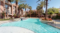 This property features a stunning resort-style pool as its centerpiece, showcasing crystal-clear turquoise water set against a backdrop of towering palm trees, lush tropical landscaping, and colorful flowering plants. The freeform pool design includes gentle water features and is framed by lounge seating areas, creating an inviting outdoor retreat beneath open blue skies. The surrounding red-brick multi-story buildings add a classic architectural character to the space.

The interior entrance area transitions seamlessly from the outdoors, featuring large-format tile flooring in a warm neutral tone with rich wood baseboards that add warmth and contrast. The entryway is appointed with a sleek wood-and-dark-finish reception console topped with a framed mirror, offering a polished and functional welcome point. Natural light filters through wood-framed glass doors, enhancing the bright, airy atmosphere.

Beyond the entry, the interior opens into a well-appointed lounge area highlighted by an eye-catching multi-arm chandelier and exposed wood ceiling details. Comfortable seating arrangements, glass-top accent tables, and curated artwork — including a large-scale abstract canvas — contribute to the space's sophisticated and welcoming ambiance throughout.