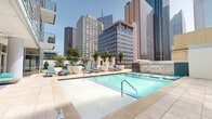 This sophisticated outdoor amenity level offers a premier urban resort experience, nestled within the heart of a bustling downtown district surrounded by soaring glass and steel skyscrapers. The centerpiece is a generously sized swimming pool with crystal-clear turquoise water, framed by elegant light stone tile decking that reflects the open sky above. An adjacent spa pool, finished with classic blue mosaic tile, provides a more intimate relaxation option alongside a signature stone water feature wall bearing the building's distinctive branding. The pool deck is thoughtfully furnished with teal-cushioned lounge chairs and seating clusters, including wooden-framed chairs and modern woven pieces, arranged to accommodate both social gatherings and quiet relaxation. A covered lounge area along one side of the deck features warm wood-paneled walls, circular decorative mirrors, and ceiling fans — offering shade and comfort without sacrificing the open-air ambiance. Potted trees and curated greenery add natural softness to the contemporary hardscape. Floor-to-ceiling glass facades of the surrounding building envelope the space, while expansive city views frame the entire experience, making this pool deck a distinctive urban retreat that seamlessly blends sophisticated design with vibrant city living.