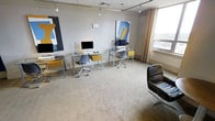 This single-floor workspace is thoughtfully designed to balance functionality with an artistic aesthetic. The open-plan layout accommodates multiple workstations, each equipped with glass-topped desks supported by sleek metal frames, desktop monitors, and ergonomic task chairs in a contemporary style. Accent yellow drawers beneath the desks add a deliberate pop of color, reinforcing the creative character of the space.

The room is finished with neutral gray carpeting and light-toned walls, providing a clean, distraction-free backdrop. Natural light enters generously through a large window fitted with sheer curtains, while a track lighting system mounted to the ceiling ensures consistent, adjustable illumination throughout the workspace.

Two bold, abstract artworks are displayed prominently on the walls — one featuring blue and yellow abstract forms and another with a striking geometric black composition — lending the space a gallery-like quality. A multicolored accent chair positioned toward the front of the room adds a casual seating option and further reinforces the eclectic, design-forward sensibility of this professional environment.