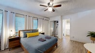 Located at 425 Second St N in the heart of downtown Saint Petersburg, Florida, this single-floor unit offers a well-organized and efficiently designed layout. The space is finished throughout with light wood-tone flooring that lends warmth and visual continuity across every room.

The main living area doubles as a bedroom, accommodating a queen-sized bed alongside a compact dining set with a round table and chairs. A wall-mounted flat-screen television is positioned for easy viewing from the sleeping area. Ceiling fans with integrated lighting fixtures are present throughout, complementing the wall-mounted mini-split air conditioning units that provide individual climate control. Natural light filters through multiple windows dressed with white grommet curtains and horizontal blinds.

The bedroom includes a built-in closet with hanging storage, and opens directly into an adjacent utility area. Walls feature a painted brick accent detail, adding a subtle architectural character to the otherwise clean, white interior.

The bathroom is finished in crisp white tile and includes a full soaking tub with a shower curtain, a pedestal sink with matte black fixtures, and a window for natural ventilation. The kitchenette features white cabinetry, light quartz-style countertops, a white electric range with a hood vent above, and a white subway tile backsplash — creating a cohesive and functional cooking space. Textured ceilings complete the interior throughout.