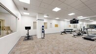 Located at 405 W Redwood St in Baltimore, MD, this fitness center offers a well-equipped, single-floor workout space designed to support a range of exercise routines. The layout is open and functional, providing ample room to move between equipment stations comfortably.

The space features a dedicated cardio zone equipped with treadmills and an elliptical trainer, all from Matrix, positioned in front of wall-mounted flat-screen TVs and a mirrored wall that enhances the sense of openness. A multi-use cable machine and functional trainer anchor the strength training area, accompanied by a rack of dumbbells and adjustable weight benches for free weight exercises. A stationary spin bike with an integrated display screen rounds out the cardio offerings, while a medicine ball rack provides options for functional fitness training.

The interior design is clean and neutral, with crisp white walls, textured loop-pile carpeting in warm beige and gray tones, and a drop ceiling fitted with recessed panel lighting that evenly illuminates the entire floor. A structural column near the center of the room adds architectural character without impeding the functional flow of the space. A bulletin board and informational signage on the walls contribute to the utility-focused atmosphere. A glass partition wall along one side of the room adds a modern, professional touch. The overall ambiance is bright, organized, and purposefully designed for fitness use.