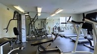 This fitness center offers a comprehensive workout environment designed to accommodate a range of exercise needs. The space is outfitted with an array of strength training equipment, including a multi-function cable machine, adjustable benches, and a leg press, alongside cardio machines such as treadmills positioned near a large window. The full-length wall mirrors amplify the sense of space and allow users to monitor their form during workouts. Overhead fluorescent lighting ensures the room is well-lit throughout, complemented by natural light streaming in through the window, which also provides views of the surrounding greenery. The flooring features dark rubber matting, a practical and durable surface suited for heavy equipment and high-traffic use. A workout reference chart posted on the wall adds a functional touch for those seeking exercise guidance. The overall layout is organized and spacious, allowing users to move freely between stations. The combination of natural light, mirrored walls, and varied equipment makes this fitness center a functional and inviting space for both strength and cardiovascular training.