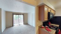 This single-floor residence offers a practical and comfortable layout designed for efficient everyday living. The living area is bright and open, featuring plush wall-to-wall carpeting, a textured ceiling, and a large sliding glass door framed by vertical blinds that opens to a private balcony, inviting natural light and outdoor views into the space. A built-in closet with louvered doors provides convenient storage near the main entry.

The galley-style kitchen is thoughtfully appointed with warm-toned wooden cabinetry offering generous storage above and below the counters. Stone-finish countertops run the length of the kitchen, complemented by a stainless steel sink, and black appliances including a refrigerator, electric range, built-in microwave, and dishwasher. Light tile flooring keeps the kitchen feeling clean and functional.

The bedroom continues the carpeted flooring found throughout the living spaces and includes a louvered closet, providing ample storage. A thermostat and HVAC venting are conveniently positioned within the room.

The bathroom features a wooden vanity with storage beneath a light countertop, a well-lit mirror flanked by a multi-bulb vanity fixture, and tile flooring. The overall palette throughout the residence is neutral and warm, creating a cohesive and inviting atmosphere.