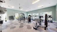 This spacious fitness center offers a well-rounded exercise environment across a single, open floor plan. The interior is finished with soft sage-green walls, white trim, and a crisp white ceiling fitted with recessed fluorescent lighting and dual ceiling fans that maintain comfortable airflow throughout the space. The distinctive flooring features a geometric hexagonal carpet tile pattern in neutral tones of gray and beige, adding visual interest underfoot.

Large floor-to-ceiling windows line multiple walls, flooding the interior with abundant natural light and providing direct views of an outdoor pool and patio area complete with umbrella-shaded seating. A set of glass double doors also connects to the exterior, reinforcing the seamless indoor-outdoor feel.

The facility is comprehensively equipped with a broad selection of strength machines, including cable systems, an Axis multi-functional trainer, seated press and curl stations, and a multi-station cable rack. Cardiovascular options include multiple treadmills and additional cardio equipment. A dedicated free weights area features tiered dumbbell racks stocked with a full range of weights, complemented by a large wall mirror that enhances the sense of space. A wall-mounted flat-screen television and ample open floor space complete this well-appointed fitness facility.