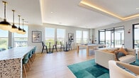 This elevated amenity lounge is a thoughtfully designed social space that seamlessly blends indoor comfort with outdoor living. The bright, open-plan interior features light hardwood-style flooring, tray ceilings with warm ambient LED cove lighting, and recessed fixtures throughout. Large windows line the walls, flooding the space with natural light and framing sweeping elevated views of the surrounding cityscape and treetops.

The interior is divided into distinct functional zones. A lounge area anchors the space with plush cream curved seating arranged around a teal geometric area rug, centered on a striking arched alcove painted in bold teal and fitted with a flat-screen TV, flanked by built-in bookshelves. Adjacent to the lounge, a pool table adds a recreational focal point. A dining and café zone features a standout terrazzo-topped communal table with patterned base, teal upholstered chairs, and pendant lighting above a tiled wet bar with open shelving.

High-top bistro tables positioned near the windows offer casual seating with panoramic views. The space flows directly onto a spacious rooftop terrace, shaded by a modern slatted pergola with string lights. The terrace includes outdoor dining tables, lounge seating, a foosball table, and an outdoor TV, all set against an open-sky backdrop with expansive city views.