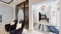 This refined single-floor space presents an exceptionally well-appointed interior that blends contemporary design with luxurious detailing throughout. The entry corridor immediately sets a distinguished tone with its bold black-and-white geometric diamond-patterned tile flooring, white structural columns with classic molding, and an eye-catching layered chandelier overhead. A large-scale framed artwork depicting draped fabric anchors one wall, complemented by plush dark velvet armchairs with cream accent pillows.

The lounge area serves as a true centerpiece, featuring a floor-to-ceiling marble-clad wall housing a sleek linear gas fireplace. A patterned sculptural ceiling, multi-toned hardwood-style flooring, and dark velvet seating arranged around a marble-topped coffee table create an intimate yet grand atmosphere.

A naturally lit seating nook with expansive floor-to-ceiling windows and sheer drapery offers an airy retreat with views of lush greenery outside. A dark wood high-top bar counter with leather stools provides an additional work or casual gathering spot.

The dining area features sophisticated booth seating with upholstered banquettes, dark tiled accent walls, and striking sculptural pendant lighting in bold yellow and gold tones, adding a vibrant artistic flourish. Throughout, large-format white tile flooring, curated abstract wall art, and cohesive dark wood accents unify the space into a polished, gallery-like environment.