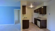This single-floor space offers a practical and straightforward layout designed for comfortable everyday living. The kitchen is a central feature, outfitted with dark wood cabinetry that provides ample storage both above and below the countertops. White appliances, including a refrigerator, electric range, and dishwasher, are seamlessly integrated into the layout. A single-basin sink is conveniently positioned beneath the upper cabinets, and tile flooring throughout the kitchen offers durability and ease of maintenance. An overhead flush-mount light fixture illuminates the space efficiently. A structural column creates a natural visual divide between the kitchen and the adjacent room, which transitions to plush carpeted flooring. This connecting room is distinguished by a vivid blue accent wall, adding a pop of color and personality to the interior. A doorway visible from this area suggests an open and accessible flow between rooms. The overall layout is compact yet well-organized, making efficient use of the available space. The combination of dark cabinetry, light countertops, and contrasting wall colors gives the interior a utilitarian character with moments of visual interest.