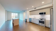 This contemporary single-floor residence showcases a clean, modern design with an open-concept layout that promotes a sense of spaciousness and effortless flow. The kitchen is a standout feature, outfitted with light wood cabinetry extending to the ceiling, a full suite of stainless steel appliances including a gas range, built-in microwave, dishwasher, and French door refrigerator, and a white subway tile backsplash. A central island with a light wood base provides additional prep space and visual continuity with the warm hardwood flooring that runs throughout the main living areas. Recessed lighting illuminates the open living zone, which benefits from large sliding glass doors and windows that invite abundant natural light and provide views of the surrounding urban landscape. The bedrooms continue the hardwood flooring and feature large windows and sliding glass doors that open to exterior balconies, enhancing the connection to the outdoors. The walk-in closet is thoughtfully designed with custom wood shelving, hanging rods, and built-in organizers set over plush carpeting. The bathroom presents a frameless glass walk-in shower enclosed in large-format neutral tile, accented with chrome fixtures and a built-in niche for a polished, spa-like finish.