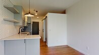This single-floor space showcases a thoughtfully designed modern interior with warm hardwood flooring running throughout. The open-concept kitchen serves as a standout feature, equipped with gray flat-front cabinetry, light quartz countertops, and a full suite of stainless steel appliances. A striking herringbone-patterned tile backsplash extends to the ceiling above the range, complemented by a statement pendant light with a copper accent. A peninsula counter provides additional prep space and casual seating, opening naturally into the living and dining area.

Sliding barn-style doors with matte black hardware add a contemporary industrial touch and help define separate areas within the layout efficiently. Track lighting in the kitchen and recessed lighting throughout provide layered illumination across the space.

The bedroom is bright and airy, benefiting from large windows that frame views of mature trees, filling the room with natural light. A sliding door leads to an adjoining space, maintaining a clean, streamlined aesthetic. The bathroom includes a full soaking tub with a shower overhead, oil-rubbed bronze fixtures, and hardwood-style flooring that ties cohesively to the rest of the unit. Overall, the space presents a clean, modern design with functional and stylish finishes throughout.