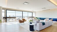 This expansive, single-floor space offers a sophisticated open-plan layout bathed in natural light, courtesy of large floor-to-ceiling windows and oversized black-framed picture windows that frame stunning panoramic views of the city skyline. Light-toned hardwood flooring runs throughout, complementing the neutral warm-beige palette of the walls and ceilings, creating a bright and airy atmosphere.

The main living area is generously proportioned, furnished with a plush sectional sofa, rounded ottoman coffee table, and an array of contemporary accent chairs arranged around a built-in wall-mounted display screen. Distinctive pendant and multi-arm chandelier lighting fixtures add a modern, sculptural quality to the space. A built-in fireplace with a bold navy blue surround serves as a striking focal point.

Adjacent to the living zone, a dedicated dining area features a substantial bar-height communal table with leather-and-metal counter stools, illuminated by a multi-arm sputnik chandelier. The open layout flows seamlessly to floor-to-ceiling sliding glass doors that lead to a spacious outdoor terrace, complete with additional seating and open-air dining. Throughout the space, carefully placed indoor greenery adds warmth and vitality, enhancing the overall inviting, contemporary ambiance.