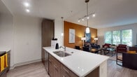 This well-designed single-floor residence offers a seamless blend of modern aesthetics and functional living. The open-concept layout connects the kitchen, dining, and living areas, creating a cohesive and inviting atmosphere. The kitchen is a standout feature, showcasing rich dark wood cabinetry, sleek white quartz countertops, a double-basin stainless steel sink set into a central island, and a full suite of stainless steel appliances including a refrigerator, microwave, and range. Pendant lighting and a track lighting system add both task illumination and visual interest overhead. Wood-look flooring runs throughout the main living areas, complementing the warm, contemporary palette. A boldly painted burnt-orange accent wall anchors the living and dining space, which benefits from natural light through generously sized windows overlooking greenery outside. The bedroom features plush carpeting, a tray ceiling with a ceiling fan, a walk-in closet, and a window that brings in additional natural light. The bathroom is finished with dark wood vanity cabinetry, white countertops, tile flooring, and a full tub-shower combination with tile surround. The overall ambiance is modern, warm, and thoughtfully designed for comfortable everyday living.