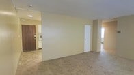 This single-floor space presents a practical and straightforward layout designed for comfortable everyday living. The main living area is finished with wall-to-wall carpeting in a neutral tone, providing a soft underfoot feel and a cohesive look throughout the space. The walls are painted in a light cream hue, contributing to a clean and understated aesthetic.

A white interior door is centrally positioned along the main wall, leading to an additional area of the unit, while a secondary doorway on the right side offers further connectivity between rooms, with a window nearby allowing natural light to filter in. The transition from the carpeted living space to the adjacent kitchen area is marked by a change in flooring, shifting to a harder surface more suitable for a cooking environment.

The kitchen is visible from the living area and features white cabinetry and appliances, maintaining the overall neutral palette of the space. A dark wood-toned entry door is also visible, providing a grounding contrast to the lighter surroundings. The ceiling is smooth and flat, fitted with recessed lighting that ensures even illumination throughout. Overall, the space offers a functional and move-in ready environment with a clean, no-frills design.