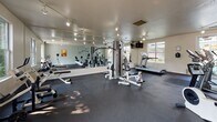 Located at 3450 El Camino Real in Santa Clara, California, this single-story fitness center offers a well-equipped space designed to support a comprehensive workout experience. The layout is thoughtfully organized to accommodate both strength training and cardiovascular exercise, providing ample room to move between stations without feeling crowded.

The strength training area is anchored by a multi-station cable machine, complemented by free weights, a dumbbell rack, and adjustable benches. A full wall of floor-to-ceiling mirrors runs along one side of the room, enhancing the sense of openness while allowing users to monitor their form. Track lighting is mounted along the ceiling, delivering consistent, directed illumination throughout the space.

The cardio section includes a treadmill, recumbent bike, and rowing machine, distributed across the room to allow comfortable use of each piece of equipment. Durable rubber flooring covers the entire space, providing both safety and stability during exercise.

Natural light enters through multiple windows, including a large sliding glass door that opens directly to an outdoor patio area, contributing to an airy, bright atmosphere. The walls are painted in neutral tones with subtle sage accents, lending a clean and calm aesthetic to the overall environment.