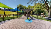 This well-appointed outdoor amenity area is designed to serve both pets and residents with a thoughtful mix of recreational features. The space is divided into distinct zones, each offering its own function within a cohesive, park-like setting.

At the entrance, a paver-lined courtyard surrounds a centrally located splash pad featuring a ground-level water play element, ideal for warm-weather enjoyment. A sturdy wooden pergola structure anchors this zone, providing partial shade over the hardscaped surface.

Adjacent to the courtyard, vibrant blue and green triangular shade sails stretch overhead, casting dappled shadows across a wide expanse of lush synthetic turf. This area includes agility equipment such as ramps and hurdles, making it well-suited for active pets. A traditional wooden play structure is also visible within a fenced enclosure nearby.

Further into the space, a more natural atmosphere prevails, with mature trees providing generous canopy coverage. Seating areas, decorative boulders, and additional agility features are scattered across open turf, offering relaxed gathering spots alongside active play.

The entire area is enclosed by black iron fencing, ensuring safety and separation between zones. Rolling green hills and a residential building complex form a scenic backdrop, completing the open, community-oriented character of this outdoor space.