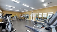 This fitness center offers a comprehensive workout environment designed to support a variety of exercise routines. The space features warm yellow walls accented by white crown molding, durable rubber flooring, and bright fluorescent lighting that ensures excellent visibility throughout. A ceiling fan contributes to comfortable air circulation during workouts. Large windows along one wall allow natural light to filter in, enhancing the overall atmosphere. The facility is well-stocked with an impressive selection of equipment, including rowing machines, adjustable weight benches, cable and pulley strength machines, and a multi-station cable system. A dedicated rack displays a colorful array of medicine balls, providing options for functional and core training. Cardio equipment, including an elliptical machine and stationary bike, is positioned near the windows. Full-length mirrors line one wall, creating a sense of expanded space while allowing users to monitor their form. A centrally mounted clock keeps members on schedule, and an informational board near the entry door provides guidance and resources. The overall layout is open and organized, offering ample room to move freely between stations and accommodate multiple users simultaneously.