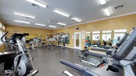This fitness center offers a comprehensive workout environment designed to support a variety of exercise routines. The space features warm yellow walls accented by white crown molding, durable rubber flooring, and bright fluorescent lighting that ensures excellent visibility throughout. A ceiling fan contributes to comfortable air circulation during workouts. Large windows along one wall allow natural light to filter in, enhancing the overall atmosphere. The facility is well-stocked with an impressive selection of equipment, including rowing machines, adjustable weight benches, cable and pulley strength machines, and a multi-station cable system. A dedicated rack displays a colorful array of medicine balls, providing options for functional and core training. Cardio equipment, including an elliptical machine and stationary bike, is positioned near the windows. Full-length mirrors line one wall, creating a sense of expanded space while allowing users to monitor their form. A centrally mounted clock keeps members on schedule, and an informational board near the entry door provides guidance and resources. The overall layout is open and organized, offering ample room to move freely between stations and accommodate multiple users simultaneously.