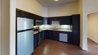This 1-bedroom, 1-bathroom unit is designed for comfortable, functional living across a single floor. The open-concept living area is anchored by rich hardwood-style flooring that extends throughout the main space, and a ceiling fan adds both style and practicality. The layout connects the living area seamlessly to the kitchen, creating an inviting, airy atmosphere.

The kitchen is a standout feature, outfitted with dark navy cabinetry that reaches toward the ceiling, providing generous storage. Granite-style countertops offer ample prep space, and the suite of stainless steel appliances — including a refrigerator, electric range, built-in microwave, and dishwasher — ensures a fully functional cooking environment. The kitchen's warm wood flooring ties it cohesively to the living space.

The bedroom is a comfortable retreat, featuring a large double-hung window that frames views of surrounding greenery and neighboring buildings, filling the room with natural light. A ceiling fan, neutral carpet, and a bold dark-panel door complete the room's clean, modern aesthetic.

The bathroom is thoughtfully finished with a dark vanity topped with granite-style countertops, a vessel-style sink, and a well-lit vanity mirror. The tub-shower combination with sliding glass doors adds a polished touch to this practical space.