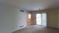 This single-floor space features a straightforward, functional layout with a focus on natural light and open living. The main living area is finished with neutral wall-to-wall carpeting and crisp white walls, creating a clean, versatile backdrop. A sliding glass door fitted with vertical blinds fills the room with natural light and offers a view of mature trees outside, providing a sense of privacy and greenery. Ceiling-mounted light fixtures and multiple floor-level and wall-mounted HVAC vents ensure year-round climate comfort throughout the space. A visible doorway connects the living area to an adjacent room, which features its own window and a ceiling fan, suggesting its use as a bedroom or secondary living space. The layout flows naturally between rooms, and the open passage between them enhances the sense of spaciousness. Overall, the space presents a neutral, move-in ready environment with practical features, ample natural light, and a calm ambiance suited for comfortable everyday living.