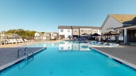 Located at 17014 Fairmount Way in Charlotte, NC, this outdoor community amenity space centers around a generously sized swimming pool with crystal-clear blue water, designed to serve as a premier gathering and recreation destination. The pool features a curved, resort-style shape with wide entry steps and ladder access, surrounded by an expansive aggregate-finish sundeck that provides ample room for lounging and socializing.

The pool deck is well-appointed with a generous arrangement of white sling-style lounge chairs, offering residents plenty of space to relax in the sun. Umbrella-shaded outdoor dining sets are also available for those seeking a cooler retreat. The area is enclosed by a sleek black wrought iron fence, providing security and a clean aesthetic boundary, with a gated entry point for controlled access.

Adjacent to the pool, a white board-and-batten clubhouse structure with pitched rooflines anchors one side of the space, offering architectural interest and complementing the multi-story residential buildings that border the community. The surrounding neighborhood features modern townhome-style architecture with gray and neutral board-and-batten siding and large windows. Under open Carolina skies, this pool area delivers a bright, welcoming outdoor environment ideal for warm-weather enjoyment.