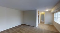 This single-floor space presents a straightforward, functional layout with clearly defined zones visible from the main living area. The primary living space is finished with wall-to-wall carpet in a neutral beige tone, providing a soft underfoot feel. The walls throughout are painted in a clean, light gray-white, contributing to a bright and open atmosphere despite the absence of visible windows in the main area.

A partial dividing wall separates the living space from a transition zone, where the flooring shifts to a light-toned tile in a geometric pattern, suggesting a kitchen or entryway area. A hallway extends from this transition zone, illuminated by a ceiling-mounted light fixture, and leads to at least one additional room visible at the far end, which also features carpeted flooring. The hallway walls maintain the same neutral palette seen throughout the space.

The ceilings are smooth and finished in white, sitting at a standard height and reinforcing the clean, unadorned aesthetic of the interior. A fire extinguisher is mounted near the hallway entrance, noting a practical safety feature. Overall, the space offers a simple, move-in-ready environment with consistent finishes across all visible areas.