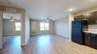 This single-floor residence offers a clean, modern layout designed for comfortable everyday living. The main living area is open and airy, featuring light wood-style luxury vinyl flooring that extends throughout the space, neutral taupe-toned walls, and white baseboards that create a bright, cohesive aesthetic. A ceiling fan with integrated lighting anchors the living room, while a window overlooking an outdoor railing admits generous natural light and outdoor views.

Adjacent to the living area, the kitchen is well-equipped with dark espresso cabinetry, light stone-look countertops, a full suite of black appliances including a refrigerator, dishwasher, range, and over-the-range microwave, recessed lighting overhead, and a sink set into an L-shaped counter configuration. The seamless transition between the kitchen and living space makes the layout feel open and connected.

The bedroom is a quiet, generously sized room with a large window framing outdoor views, a built-in closet with rod and shelf storage, and the same consistent flooring found throughout. A utility access panel is also present within the living area. The bathroom features large-format white tile flooring, a full soaking tub with shower, a toilet, and a vanity with dark cabinet storage, offering a clean and functional design.