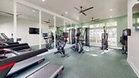 Located in North Charleston, SC, this fitness center offers a thoughtfully designed exercise environment that accommodates a wide range of workout styles. The space is finished with durable, green rubber flooring throughout, providing both comfort and functionality underfoot. Recessed lighting and multiple ceiling fans ensure the room remains bright and well-ventilated during any workout session.

Large windows along one wall flood the interior with generous natural light and offer views of the surrounding greenery, creating an energizing atmosphere. Full-length framed mirrors line key sections of the walls, visually expanding the space and allowing exercisers to monitor their form.

The facility is comprehensively equipped with cardio machines including spin bikes, an elliptical, a stair climber, and a treadmill, as well as a multi-station cable machine and additional strength training equipment. Free weights, medicine balls, battle ropes, and a plyo box round out the functional fitness offerings. Wall-mounted TVs provide entertainment options throughout the workout.

A built-in shelving unit with a beverage refrigerator and counter space offers a convenient amenity station, while a dedicated storage area keeps accessories organized. A glass-paneled door provides direct outdoor access, further enhancing the open, airy character of this well-appointed fitness facility.