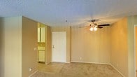 This single-floor residential space offers a practical and functional layout designed for comfortable everyday living. The main living area is finished with wall-to-wall carpeting that adds warmth underfoot, while a ceiling fan with an integrated light fixture provides both airflow and ambient lighting. The textured, popcorn-style ceiling is a notable architectural detail found throughout the space. Neutral-toned walls create a clean and versatile backdrop suitable for a variety of interior styles. A white paneled door serves as the primary entry point to the space, lending a classic and tidy appearance. Adjacent to the living area, the kitchen is visible through an open pass-through, revealing white cabinetry, countertop space, and standard appliances, including what appears to be a refrigerator and dishwasher. The kitchen's layout is efficient and well-organized, making it easy to utilize. The overall ambiance of the space is bright, understated, and move-in ready, with a cohesive color palette of warm beiges and whites that tie the rooms together seamlessly.