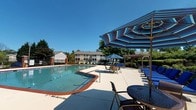 This swimming pool facility offers a well-designed outdoor recreation area complemented by an attached amenity building. The expansive pool features a brick-trimmed surround and clear, blue water, with lane markers indicating designated swim areas and posted depth signage. The surrounding concrete deck is generously sized, accommodating multiple seating arrangements including loungers, tables, and chairs shaded by large blue-and-white striped umbrellas. A pergola structure anchors one end of the deck, while mature trees and black iron perimeter fencing frame the space. The building adjacent to the pool provides direct pool access through a windowed exterior door. Inside, the corridors are finished with large-format dark tile flooring and neutral gray walls, creating a clean, modern aesthetic. A glass-walled fitness center is visible from the hallway, equipped with cardio machines and a wall-mounted television. The building also includes a restroom, dual stainless steel wall-mounted water fountains, and a staircase with carpeted treads leading to additional levels. A glimpse into an adjacent carpeted common room suggests additional gathering space nearby. Overall, the facility is well-maintained and designed to serve as a functional, community-oriented amenity.
