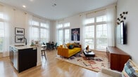 Located at 200 E Cama St in Charlotte, NC, this thoughtfully designed single-floor residence combines modern aesthetics with a warm, eclectic sensibility. Light hardwood flooring flows seamlessly throughout, anchoring each space with a clean and cohesive foundation.

The open-concept living and dining area is a standout, bathed in natural light streaming through oversized windows dressed with sheer white curtains and wood blinds. The living room features soaring ceilings, bold artwork, and a vibrant yellow sofa set atop a patterned area rug, creating an inviting and energetic atmosphere. A dedicated dining nook sits beside floor-to-ceiling windows, offering an airy setting for meals.

The kitchen is sleekly appointed with two-tone cabinetry—white upper cabinets and dark lower cabinets—complemented by stainless steel appliances, a tile backsplash, and a breakfast bar. An adjacent workspace provides a compact and functional area for remote work or study.

The bedroom offers a serene retreat, highlighted by a striking geometric accent wall, large windows with outdoor views, and ample storage. A well-designed hallway connects the living areas to the private spaces, maintaining a smooth and logical flow.

The bathroom is finished with a broad white vanity, gold-toned hardware, a walk-in glass shower, and contemporary pendant lighting, delivering a clean and modern aesthetic.