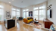 Located at 200 E Cama St in Charlotte, NC, this thoughtfully designed single-floor residence combines modern aesthetics with a warm, eclectic sensibility. Light hardwood flooring flows seamlessly throughout, anchoring each space with a clean and cohesive foundation.

The open-concept living and dining area is a standout, bathed in natural light streaming through oversized windows dressed with sheer white curtains and wood blinds. The living room features soaring ceilings, bold artwork, and a vibrant yellow sofa set atop a patterned area rug, creating an inviting and energetic atmosphere. A dedicated dining nook sits beside floor-to-ceiling windows, offering an airy setting for meals.

The kitchen is sleekly appointed with two-tone cabinetry—white upper cabinets and dark lower cabinets—complemented by stainless steel appliances, a tile backsplash, and a breakfast bar. An adjacent workspace provides a compact and functional area for remote work or study.

The bedroom offers a serene retreat, highlighted by a striking geometric accent wall, large windows with outdoor views, and ample storage. A well-designed hallway connects the living areas to the private spaces, maintaining a smooth and logical flow.

The bathroom is finished with a broad white vanity, gold-toned hardware, a walk-in glass shower, and contemporary pendant lighting, delivering a clean and modern aesthetic.