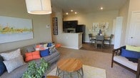 This single-floor unit showcases an open-concept floor plan that seamlessly integrates the living, dining, and kitchen areas into one cohesive, functional space. The living area features plush carpeting and is furnished with a comfortable sofa adorned with colorful accent pillows, complemented by a round wooden coffee table that adds a warm, contemporary touch. Recessed lighting throughout provides consistent, ambient illumination across the space. The kitchen is positioned centrally within the open layout, featuring dark cabinetry, black appliances including a refrigerator and microwave, and a breakfast bar that serves as a natural divider between the kitchen and living areas. The breakfast bar offers additional seating and counter space, enhancing the kitchen's practicality. Adjacent to the kitchen, the dining area accommodates a mid-sized table with upholstered chairs, making it suitable for everyday meals or entertaining. Neutral-toned walls throughout create a versatile backdrop, while decorative wall art adds personality to the space. The combination of carpet in the living area and hard-surface flooring near the kitchen and dining zones provides both comfort and easy maintenance.