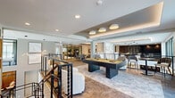 This expansive amenity lounge is a thoughtfully designed, multi-zone social space that blends entertainment, comfort, and contemporary style across a single generously sized floor. The main lounge area serves as the centerpiece, featuring a full-size pool table set atop a herringbone-patterned area rug, surrounded by curved swivel lounge chairs and accented by a coffered ceiling with drum pendant lighting. A dedicated games area offers a poker/card table with stylish houndstooth chairs, a foosball table, and an air hockey table positioned beneath a distinctive olive green ceiling with warm globe fixtures. Floor-to-ceiling windows and glass sliding doors flood the space with natural light and provide outdoor views throughout. A sleek bar counter with leather stools creates a social focal point, while a vibrant kitchenette features stainless steel appliances, a colorful herringbone tile backsplash, and bold yellow accent walls. A glass-enclosed fitness studio is visible from the main lounge, offering a seamless connection between active and social areas. Marble-effect stone feature walls, curated artwork, open cable railings, and warm wood-tone flooring complete the polished, resort-inspired ambiance of this versatile communal space.