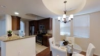 This single-floor residence showcases a thoughtfully designed open-concept layout connecting the kitchen and dining area in a seamless, functional arrangement. The kitchen is outfitted with rich, dark-stained wood cabinetry that extends to the ceiling, providing ample storage space. Granite countertops add a refined touch, while stainless steel appliances — including a gas range, microwave, and refrigerator — offer a modern, cohesive aesthetic. A tiled backsplash and light tile flooring complete the kitchen's polished look. A peninsula with a granite countertop serves as a natural divider between the kitchen and adjacent spaces, adding convenience for casual dining or meal prep. The dining area features warm hardwood flooring and is anchored by a circular glass-top table surrounded by white upholstered chairs, creating an inviting atmosphere. A wrought-iron chandelier with frosted glass shades hangs above the table, providing elegant ambient lighting. Natural light fills the dining space through a large window dressed with horizontal blinds. The interplay of dark cabinetry, warm wood tones, and neutral walls creates a balanced, welcoming ambiance throughout.