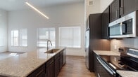This single-story residence is defined by its striking soaring ceilings, which lend an expansive, airy quality throughout the open-concept living and kitchen areas. The kitchen serves as the focal point of the main living space, anchored by a large central island topped with light granite countertops that also features an undermount sink and built-in dishwasher. Rich, dark cabinetry lines the walls and island, offering generous storage, while a classic tile backsplash adds texture behind the cooking area. Stainless steel appliances — including a French door refrigerator, electric range, and over-the-range microwave — complete the modern kitchen suite. Wood-look plank flooring runs throughout the main living areas, complementing the warm, contemporary aesthetic. Large windows fitted with horizontal blinds invite abundant natural light into both the kitchen and adjoining living space. A ceiling fan is present in the living area, enhancing comfort and airflow beneath the tall ceilings. A linear pendant light fixture provides focused illumination over the kitchen island. The bedroom is carpeted and well-lit by a sizable window, offering a quiet, neutral retreat. A spacious walk-in closet with built-in shelving and hanging rods provides excellent storage. The overall ambiance is clean, modern, and bright.