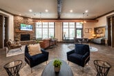 This thoughtfully designed single-story clubhouse blends modern comfort with rustic charm through a cohesive architectural language of exposed wooden beams, copper ductwork, natural stone accents, and large multi-pane windows that flood the interior with natural light. The expansive main lounge serves as the heart of the space, anchored by a floor-to-ceiling stacked stone fireplace with a mounted television above, and furnished with a mix of cognac leather sectionals, navy blue accent chairs, and coffee tables arranged across stone tile flooring. Multiple seating vignettes accommodate both social gatherings and quiet relaxation, while a casual dining area with bistro-style tables sits nearby. A striking living wall of lush greenery adds a vibrant, organic element near the main entrance. The leasing and administrative area features private offices with large interior windows overlooking the lounge, warm wood desks, and plush carpeted floors. A well-appointed kitchen includes rich wood cabinetry, granite countertops, stainless steel appliances, and a mosaic tile backsplash. The fitness center is generously equipped with cardio machines, cable strength equipment, free weights, stability balls, and a water station — all bathed in natural light from surrounding windows.