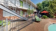 Located at 1610 Allen Toussaint Blvd in New Orleans, LA, this outdoor courtyard space captures the distinctive character and charm of the Crescent City. At its centerpiece is a generously sized freeform swimming pool with striking turquoise water, surrounded by a spacious pool deck appointed with lounge chairs, a hot tub, and shaded seating areas with red umbrellas — ideal for relaxation in the warm Louisiana climate.

The surrounding building showcases classic New Orleans architectural details, including exposed brick facades, bold lime-green accent doors, and ornate wrought-iron balcony railings that span the upper level. Decorative ironwork columns add an additional layer of craftsmanship and regional character to the structure. Stairways connecting the ground level to the upper balconies are framed with matching ironwork, reinforcing the cohesive aesthetic throughout.

Tropical landscaping, including tall palm trees and lush greenery, frames the courtyard and enhances the resort-like atmosphere. A charming brick poolhouse with a sloped roof and vibrant green door serves as a functional and decorative focal point along the pool's edge. The overall ambiance is lively yet inviting, blending New Orleans cultural flair with comfortable outdoor living in a well-maintained, enclosed courtyard setting.