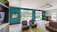 Located at 4915 Misty Oaks Dr in Charlotte, NC, this single-floor community clubhouse is thoughtfully designed to serve as a social and recreational hub. The space immediately stands out with its bold teal accent walls and large glass garage-style doors that open the interior to lush outdoor views, allowing abundant natural light to pour throughout.

The main lounge area is anchored by a plush brown sofa and coordinating armchairs accented with teal and navy throw pillows, all arranged around a white coffee table atop a green area rug. A full-length shuffleboard table provides a central entertainment focal point, complemented by a built-in media wall featuring dark cabinetry, open shelving with greenery, and dual flat-screen televisions — ideal for gatherings and game days.

An adjacent dining area offers a dark-topped table surrounded by vibrant yellow chairs, positioned near floor-to-ceiling glass French doors that provide direct sightlines to the outdoor pool area. Light tile flooring flows seamlessly throughout, tying together each zone.

Abstract, colorful artwork displayed throughout the space adds a curated, contemporary feel, enhancing the overall modern aesthetic. The combination of indoor-outdoor connectivity, entertainment amenities, and stylish furnishings makes this clubhouse an inviting retreat for residents of the Misty Oaks community.