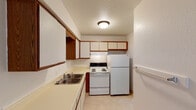 This single-floor unit offers a practical and straightforward layout with clearly defined living spaces. The kitchen is efficiently arranged in an L-shaped configuration, featuring white upper cabinets with warm wood-tone trim, a double stainless steel sink set into a light-colored countertop, and white appliances including a coil-burner electric range and a full-size refrigerator. Light tile flooring and a textured ceiling with a flush-mount light fixture complete the kitchen's functional character.

The living area is open and spacious, with neutral beige carpeting underfoot and white walls that create a clean, versatile backdrop. A wall-mounted air conditioning unit and baseboard heating provide year-round climate control. The layout, visible in the 3D model, shows a connected flow between the living space and the kitchen.

The bedrooms continue the neutral palette with beige carpet, white walls, textured ceilings, and baseboard heaters. Natural light enters through windows fitted with horizontal blinds, and closet access is provided within the rooms. Crown molding adds a subtle architectural detail.

The bathroom includes a single-sink vanity with a large mirror, a combination tub and shower unit, tile flooring, and a full vanity light bar above the mirror, offering a clean and functional design.