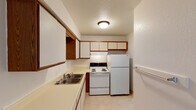 This single-floor unit offers a practical and straightforward layout with clearly defined living spaces. The kitchen is efficiently arranged in an L-shaped configuration, featuring white upper cabinets with warm wood-tone trim, a double stainless steel sink set into a light-colored countertop, and white appliances including a coil-burner electric range and a full-size refrigerator. Light tile flooring and a textured ceiling with a flush-mount light fixture complete the kitchen's functional character.

The living area is open and spacious, with neutral beige carpeting underfoot and white walls that create a clean, versatile backdrop. A wall-mounted air conditioning unit and baseboard heating provide year-round climate control. The layout, visible in the 3D model, shows a connected flow between the living space and the kitchen.

The bedrooms continue the neutral palette with beige carpet, white walls, textured ceilings, and baseboard heaters. Natural light enters through windows fitted with horizontal blinds, and closet access is provided within the rooms. Crown molding adds a subtle architectural detail.

The bathroom includes a single-sink vanity with a large mirror, a combination tub and shower unit, tile flooring, and a full vanity light bar above the mirror, offering a clean and functional design.