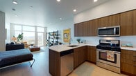 This single-floor space showcases a thoughtfully designed open-concept layout with contemporary finishes throughout. The kitchen is a standout feature, equipped with warm walnut-toned cabinetry extending to the ceiling, crisp white quartz countertops, a peninsula with an undermount sink, and a full suite of stainless steel appliances including a gas range, microwave, dishwasher, and refrigerator. Light hardwood flooring runs continuously through the kitchen into the living area, enhancing the sense of openness.

The living area benefits from dramatic floor-to-ceiling windows that flood the space with natural light and offer panoramic outdoor views. Recessed lighting complements the bright, airy atmosphere created by the white walls and high ceilings.

The bathroom features a glass-enclosed walk-in shower with mosaic tile flooring and clean white tile walls, maintaining a minimalist aesthetic. A stacked Whirlpool washer and dryer unit is conveniently integrated within the bathroom area, maximizing space efficiency. A separate water closet provides additional privacy. The overall ambiance is modern, clean, and functional, with cohesive design elements tying each area together seamlessly.